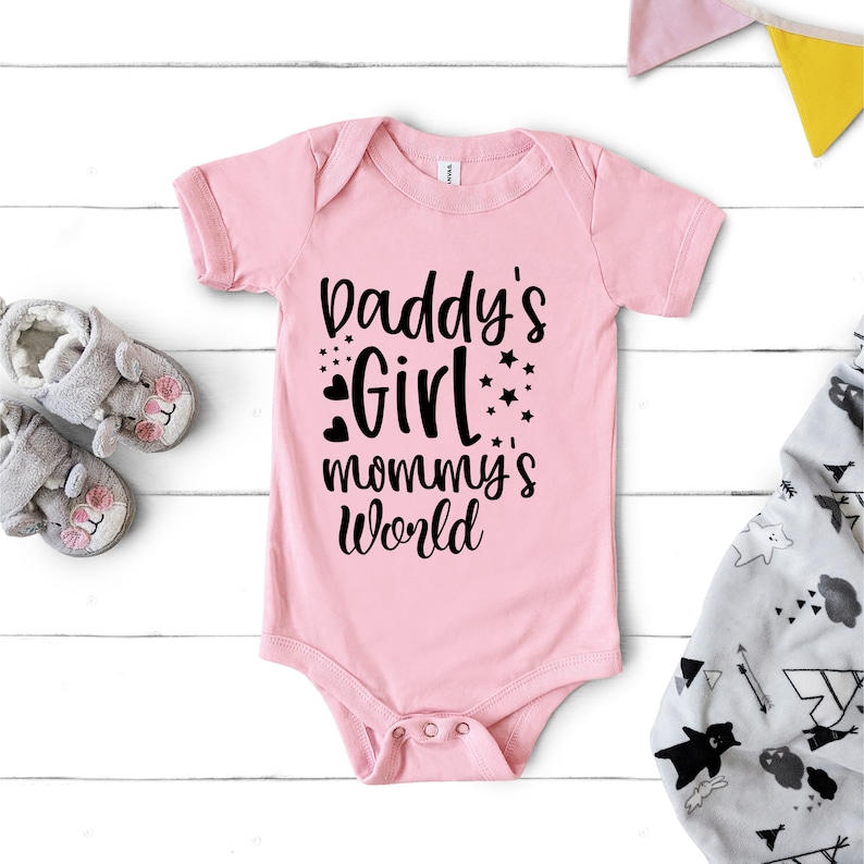 Puede incluir: Un body de beb&eacute; rosa con texto negro que dice "Daddy's Girl mommy's world" con peque&ntilde;os corazones y estrellas alrededor del texto.