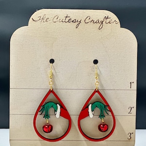 May include: Red teardrop-shaped earrings with a green hand holding a red bell. The earrings are approximately 2 inches long. The earrings are made of wood and have a gold-colored hook.