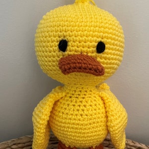 May include: A crocheted yellow duck toy with orange feet. The duck has black eyes and a brown beak.