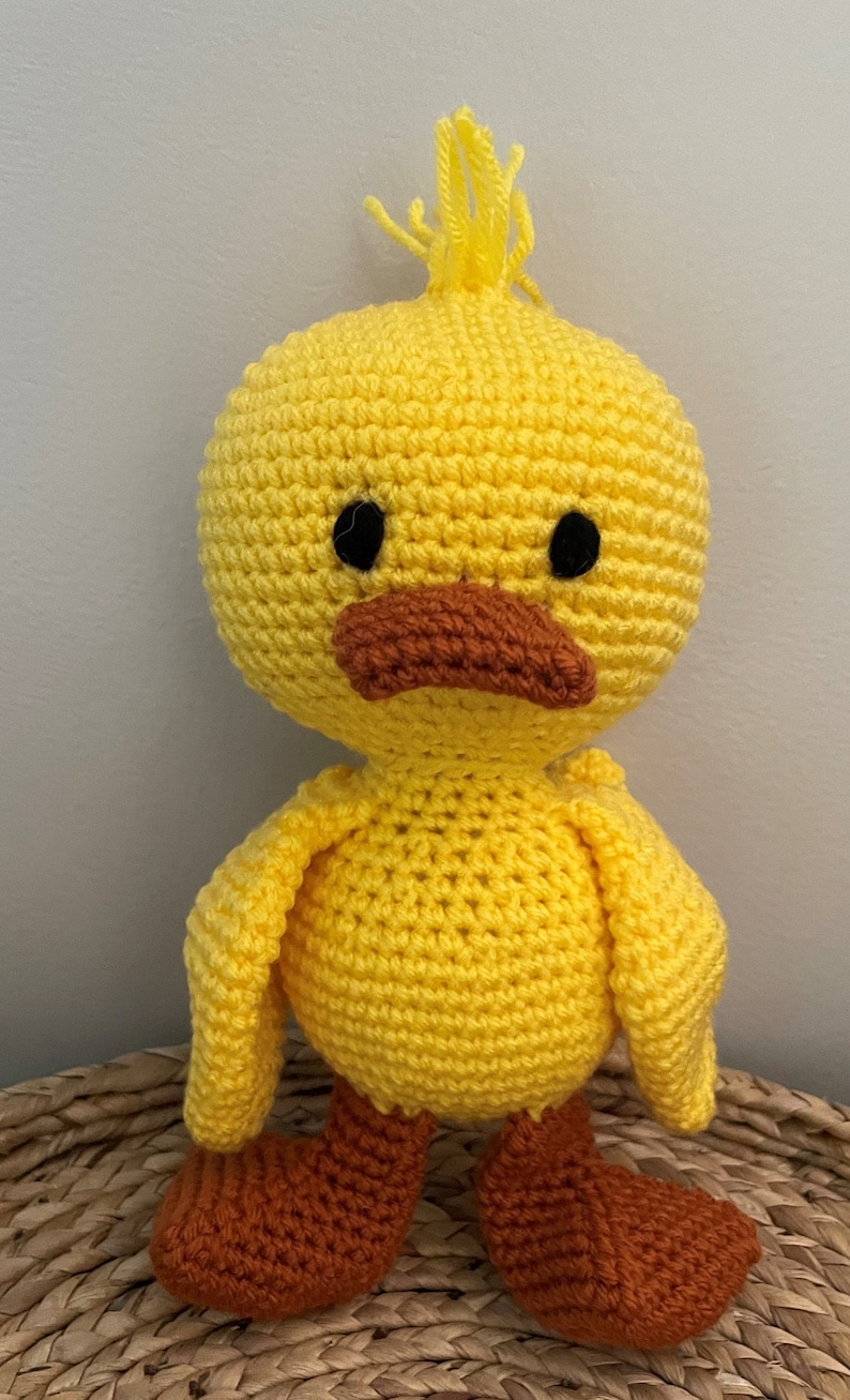 May include: A crocheted yellow duck toy with orange feet. The duck has black eyes and a brown beak.