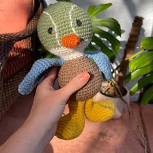 May include: A crocheted stuffed duck toy with green, brown, blue, yellow, and orange accents. The duck has a long orange beak and black eyes.