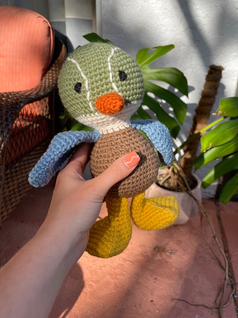 May include: A crocheted stuffed duck toy with green, brown, blue, yellow, and orange accents. The duck has a long orange beak and black eyes.