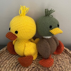 May include: Two crocheted stuffed animals, a yellow duck and a green duck, sitting on a woven basket. The yellow duck has orange feet and a black eye. The green duck has brown feet and a black eye.