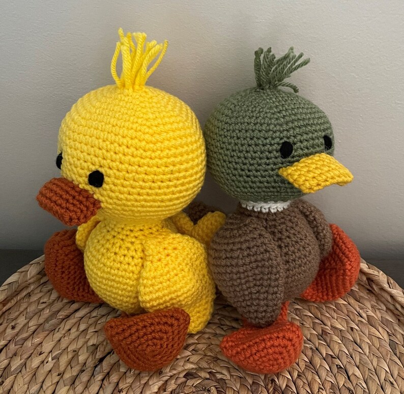May include: Two crocheted stuffed animals, a yellow duck and a green duck, sitting on a woven basket. The yellow duck has orange feet and a black eye. The green duck has brown feet and a black eye.