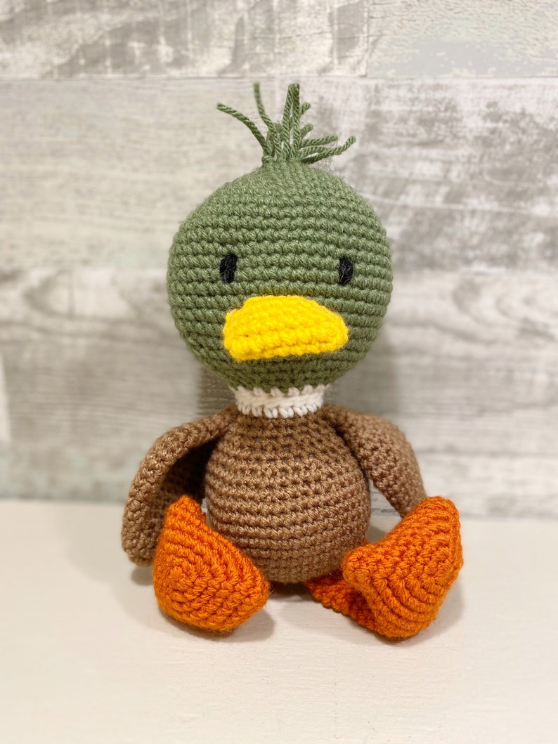 May include: A crocheted duck toy with green and brown body, yellow beak, and orange feet.