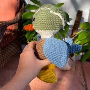 May include: A crocheted stuffed duck toy with a green head, brown body, blue wings, and yellow feet. The duck is wearing a white crocheted hat with a green brim.