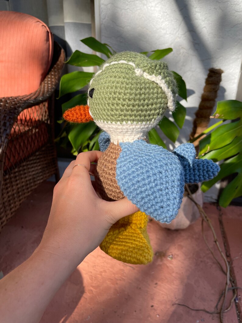May include: A crocheted stuffed duck toy with a green head, brown body, blue wings, and yellow feet. The duck is wearing a white crocheted hat with a green brim.