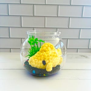 May include: A clear glass fishbowl with a yellow crocheted shark, green plastic plants, and black and colorful gravel. The shark has a black eye and is resting on the gravel. The background is a white brick wall.