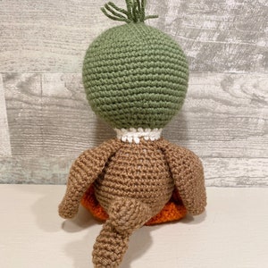 May include: A crocheted duck plush toy with a green head, brown body, and orange feet. The duck is sitting on its tail.