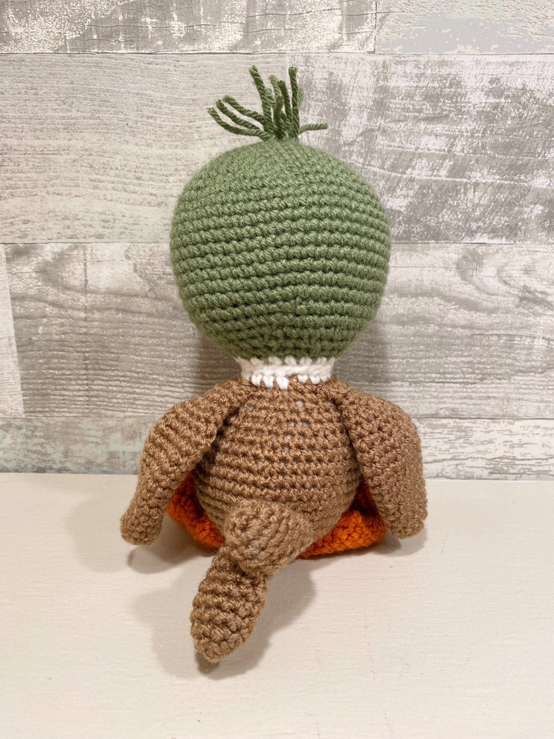 May include: A crocheted duck plush toy with a green head, brown body, and orange feet. The duck is sitting on its tail.
