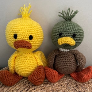 May include: Two crocheted stuffed animals, a yellow duck and a green duck, are sitting on a woven basket. The yellow duck has a brown beak and black eyes. The green duck has a brown beak and black eyes.