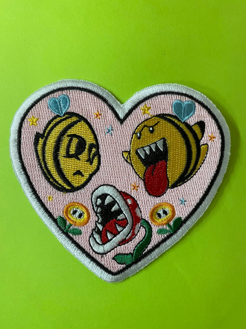 May include: A pink heart-shaped patch with two yellow and black bees, one with a smiling face and the other with a toothy grin and a red tongue. The bees are surrounded by small white stars and yellow flowers. At the bottom of the patch is a green plant with a red and white flower with a large open mouth.