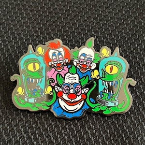 May include: A metal pin featuring four cartoon characters. The characters are a green monster with one eye, a pink and white clown with a red nose, a blue and white clown with a red nose, and a green and white clown with a red nose. The pin is a fun and quirky addition to any collection.