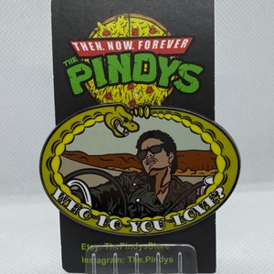 May include: A yellow and silver enamel pin featuring a cartoon image of a man driving a car with a snake in the background. The pin has the text "Who Do You Love?" around the edge. The pin is on a black card with the text "The Pindys" in green and yellow.