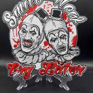 May include: Embroidered patch featuring two sinister clown faces with top hats, set against a red-splattered white background. The patch reads "Smile Now, Cry Later" in a stylized font, perfect for jackets, bags, or other accessories.
