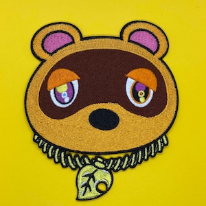 May include: Embroidered patch of Tom Nook from the Animal Crossing video game series. The patch features Tom Nook's face with his signature brown nose and large eyes. He is wearing a yellow and brown shirt with a gold chain and a green leaf pendant.