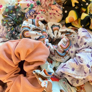 May include: A collection of colorful fabric scrunchies with various patterns, including polka dots, rainbows, and floral designs. The scrunchies are made of soft, stretchy fabric and are perfect for holding hair in place.