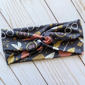 May include: A dark gray headband with a repeating pattern of white glasses, gold lightning bolts, and pink and gold illustrations. The headband is knotted at the top.