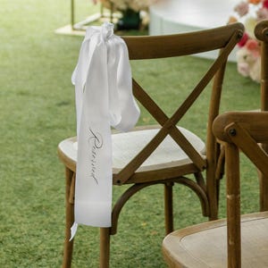May include: Outdoor wedding ceremony setup with wooden chairs on green grass. A white ribbon with the word "Reserved" hangs from a chair. Floral arrangements in shades of pink, white, and red decorate the background.