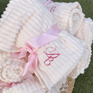 Puede incluir: Una pila de mantas suaves de color crema atadas con una cinta de raso rosa. Las mantas tienen una textura acanalada y están bordadas con un monograma rosa. Las mantas se presentan en una cesta de mimbre sobre una superficie verde.