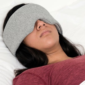 May include: A person wearing a gray sleep mask, lying on a white bed. The mask covers the eyes and forehead. The person is wearing a red long-sleeve shirt. The image is well-lit and the focus is on the mask.