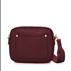 May include: A burgundy crossbody bag with a textured finish. The bag features a rectangular shape with a zippered top, a front pocket, and gold-colored hardware. It has a matching adjustable strap.
