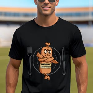 Può includere: T-shirt nero con un'illustrazione di salsiccia dei cartoni animati. La salsiccia ha le braccia incrociate e un'espressione arrabbiata, su un piatto con forchetta e coltello. L'etichetta dice "beleidigte Leberwurst".