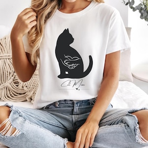 Könnte beinhalten: Weißes T-Shirt mit einer schwarzen Katzen-Silhouette, die ein Herz hält, und dem handgeschriebenen Text "Cat Mom". Das Design enthält zwei kleine Herzsymbole. Das T-Shirt ist aus einem weichen Material gefertigt.