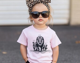 May include: A light pink t-shirt featuring a black and white lion family graphic. The graphic includes a large lion and two smaller lion cubs. The child is wearing a leopard print headband and sunglasses.
