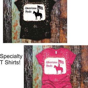 May include: Two bleached t-shirts with a distressed look. The shirts feature a silhouette of a woman riding a horse with an American flag. The text "American Made" is printed on the shirts.