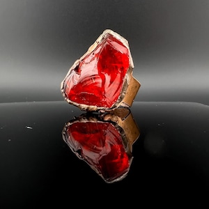 May include: A red glass ring with a brass setting. The ring is shaped like a large, irregular piece of glass.
