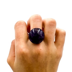 Può includere: Un anello in ametista viola con taglio cabochon rotondo. L'anello è indossato su un dito.