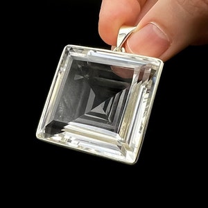 May include: A clear, square-cut pendant with a silver bail. The pendant has a multi-faceted design, creating a geometric pattern within the transparent crystal. The background is solid black, highlighting the pendant's clarity and shape.