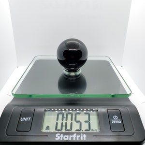 Op de afbeelding: Een zwarte glazen bol op een digitale weegschaal die 0,053 aangeeft. De weegschaal is zwart met een glazen platform en een wit digitaal display.