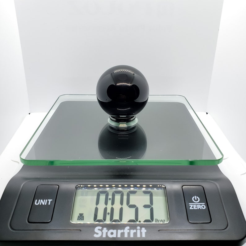 Op de afbeelding: Een zwarte glazen bol op een digitale weegschaal die 0,053 aangeeft. De weegschaal is zwart met een glazen platform en een wit digitaal display.