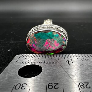 May include: A large, oval-shaped ring with a vibrant, multi-colored gemstone. The stone displays shades of green, pink, and purple. The ring has a silver band with intricate detailing. A ruler is placed next to the ring, showing measurements in inches.