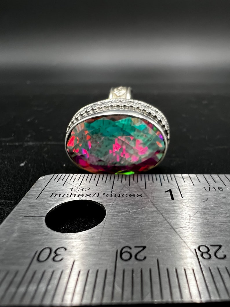 May include: A large, oval-shaped ring with a vibrant, multi-colored gemstone. The stone displays shades of green, pink, and purple. The ring has a silver band with intricate detailing. A ruler is placed next to the ring, showing measurements in inches.