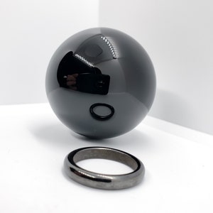 Op de afbeelding: Een zwarte obsidiaankogel en een zwarte obsidiaanring. De bol is hoogglanz gepolijst en reflecteert licht. De ring is eenvoudig en heeft een gladde, gepolijste afwerking.