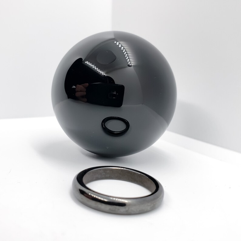 Op de afbeelding: Een zwarte obsidiaankogel en een zwarte obsidiaanring. De bol is hoogglanz gepolijst en reflecteert licht. De ring is eenvoudig en heeft een gladde, gepolijste afwerking.