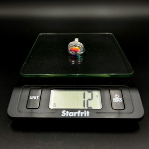 May include: A silver pendant with a colorful, iridescent stone is displayed on a digital scale. The scale reads 12 grams. The pendant has a detailed, ornate setting.