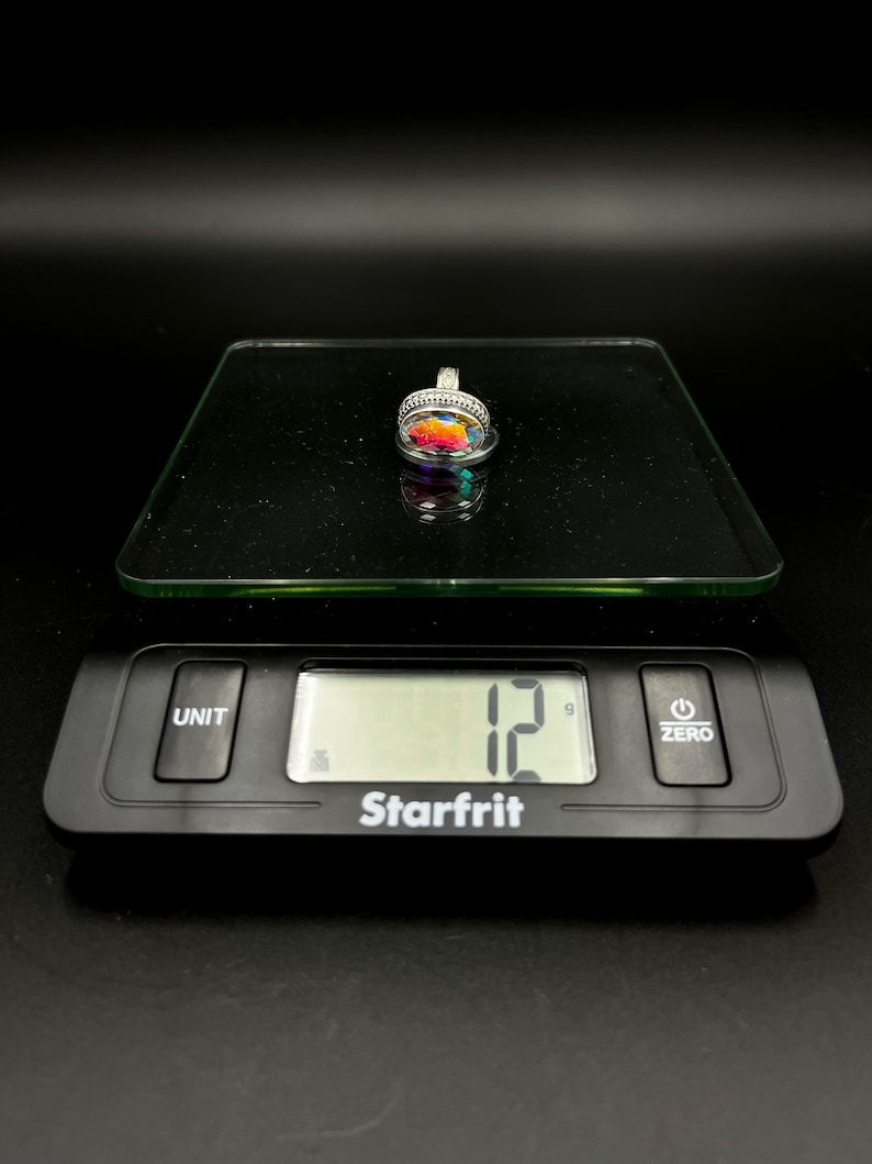 May include: A silver pendant with a colorful, iridescent stone is displayed on a digital scale. The scale reads 12 grams. The pendant has a detailed, ornate setting.