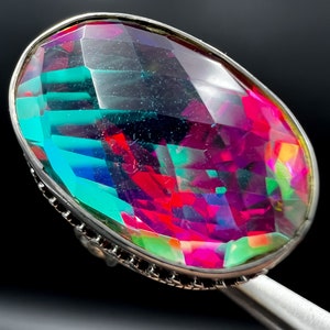 May include: A large, oval-shaped ring with a faceted, multi-colored gemstone. The gem displays vibrant hues of teal, magenta, and green, set in a silver-toned metal band. The ring is held by a silver-colored tool against a black background.