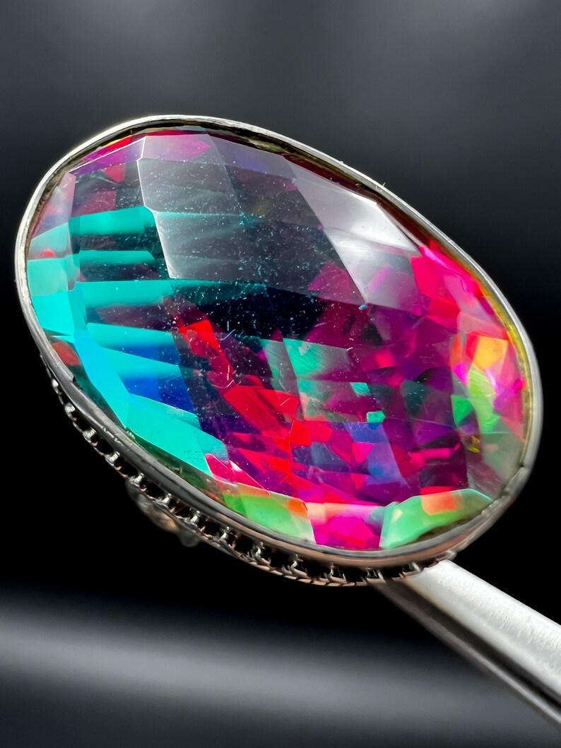 May include: A large, oval-shaped ring with a faceted, multi-colored gemstone. The gem displays vibrant hues of teal, magenta, and green, set in a silver-toned metal band. The ring is held by a silver-colored tool against a black background.