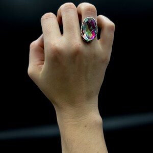 May include: A large, oval-shaped ring with a faceted, iridescent gemstone. The gemstone displays a spectrum of colors, including pink, green, and purple. The ring has a silver-colored band with a detailed, textured design around the gemstone.