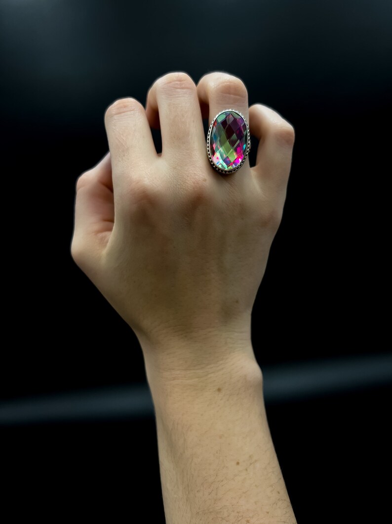May include: A large, oval-shaped ring with a faceted, iridescent gemstone. The gemstone displays a spectrum of colors, including pink, green, and purple. The ring has a silver-colored band with a detailed, textured design around the gemstone.