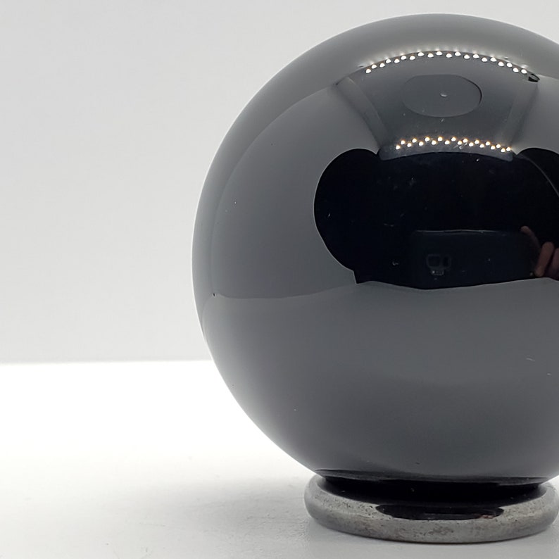 Op de afbeelding: Een zwarte glazen bol op een zilveren standaard. De bol is zeer reflecterend en lijkt van gepolijst obsidiaan te zijn.
