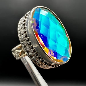 May include: A large, oval-cut gemstone ring with a silver band. The gemstone displays a vibrant, iridescent color palette of blues, greens, and yellows. The ring's band features intricate, decorative detailing.