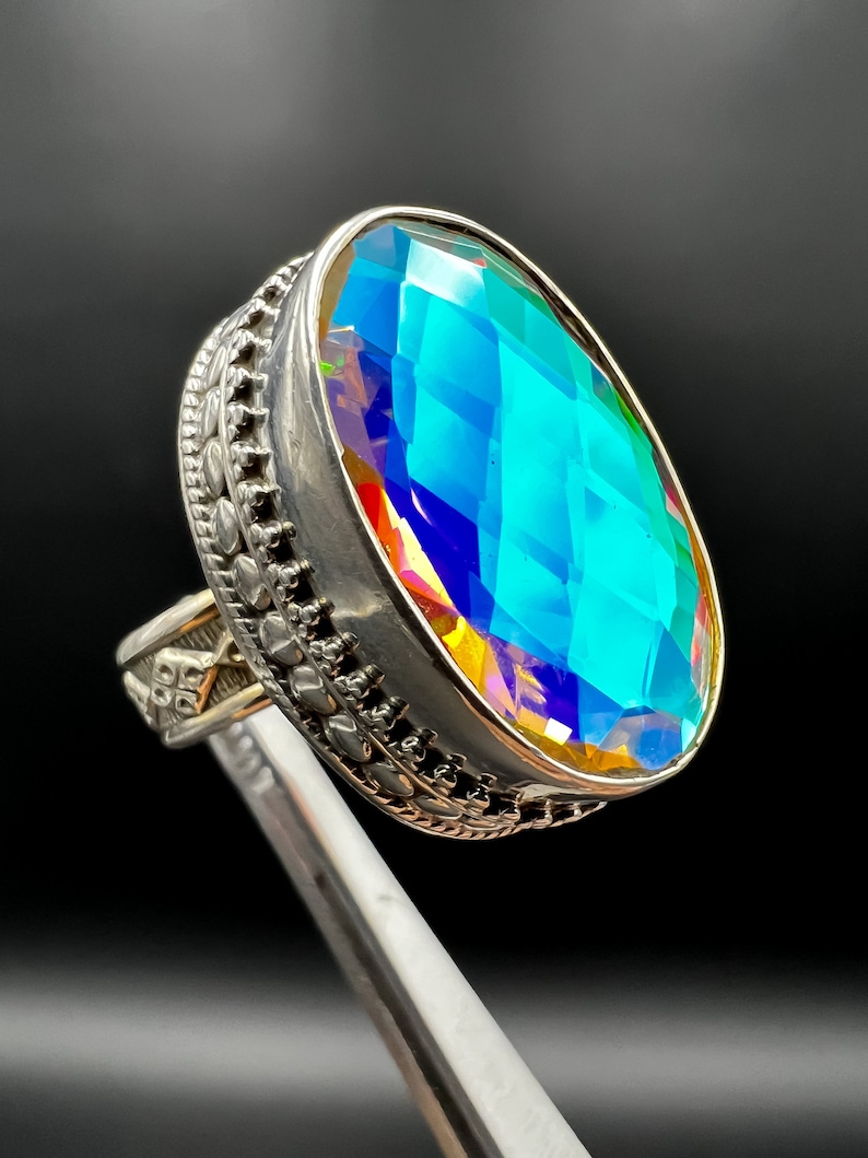 May include: A large, oval-cut gemstone ring with a silver band. The gemstone displays a vibrant, iridescent color palette of blues, greens, and yellows. The ring's band features intricate, decorative detailing.