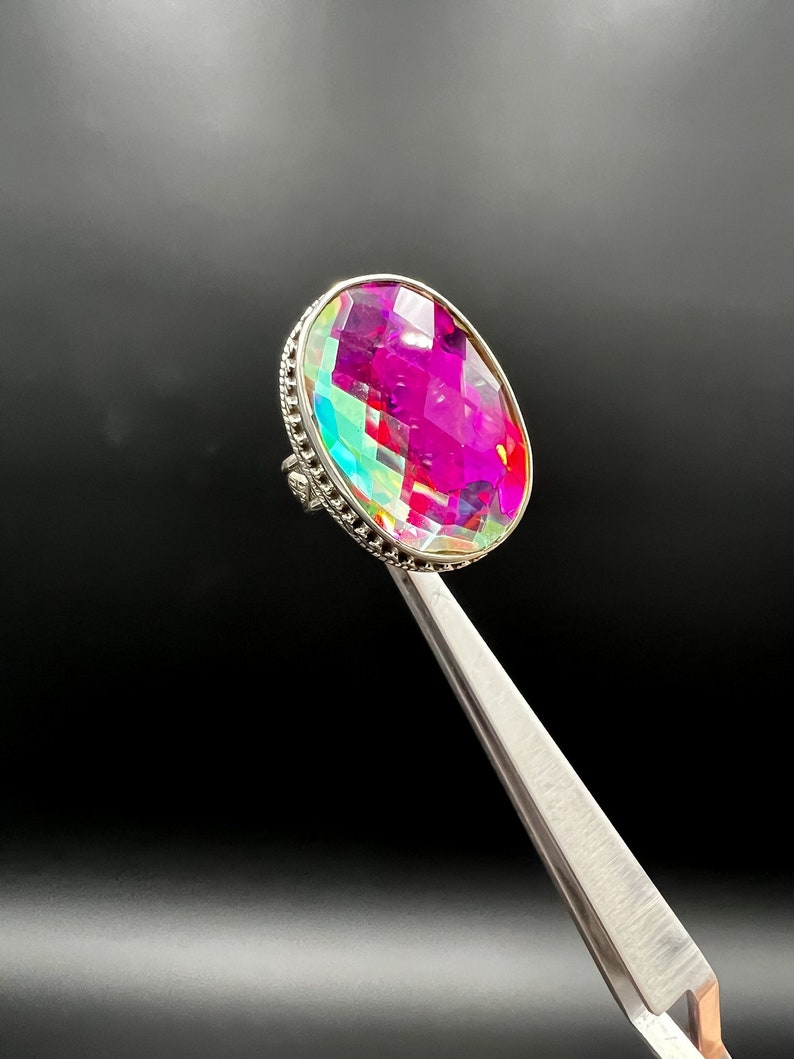 May include: A silver ring featuring a large, oval-cut gemstone with a vibrant display of pink, purple, and green hues. The gemstone is held by a detailed silver setting. The ring is held by a pair of tweezers against a black background.