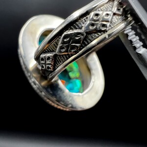 May include: Close-up of a silver ring with an oval-shaped, iridescent gemstone. The ring's band features a detailed, geometric pattern. The gemstone displays a vibrant play of colors, including blues, greens, and oranges, against a dark background.